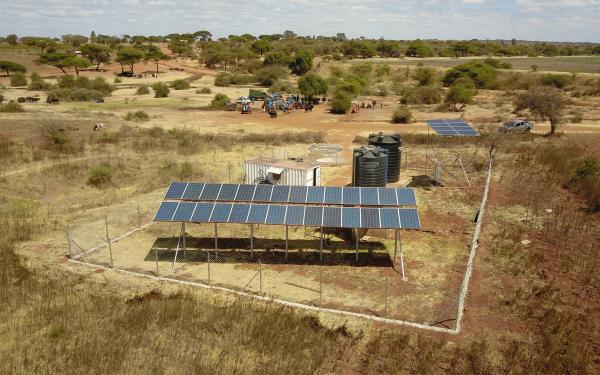 2 sets of solar panels perched up on metal frames fenced in with wire fence with 2 large black water cylindrical water reservoirs and a white trailer surrounded by dirt, dry grass and trees with people and animals in a distance. 