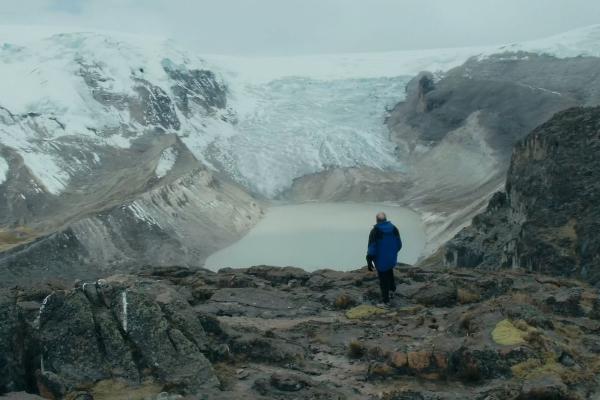 A man standing on top of a rocky mountain facing a glacier and a lake in front of it, in a valley with snow covering the surrounding mountains.