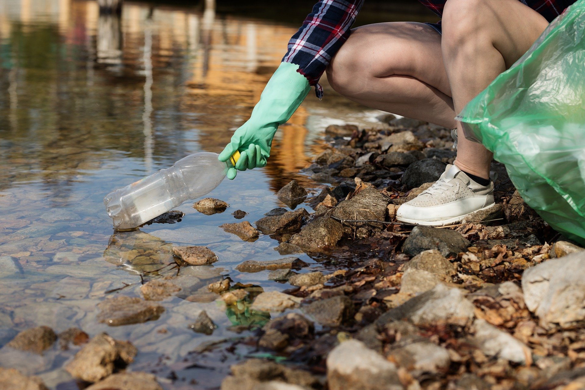 images shows legs of someone knelt down taking out a bottle from the edge of a stream wearing shorts, sneakers, green rubber gloves and a trashbag