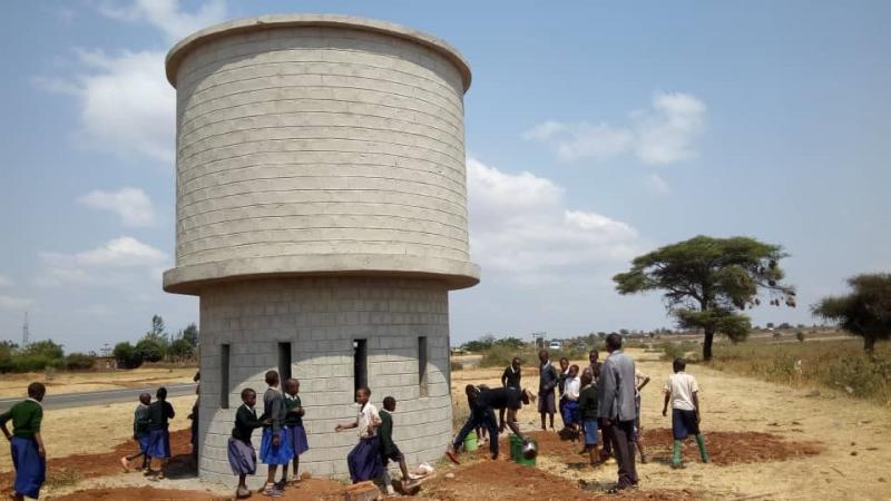 under blue skies and white clouds, some trees in a distance and dirt and hay covered ground A number of kids and adults standing around what looks like a two-story circular cement structure with long rectangular windows on first floor.