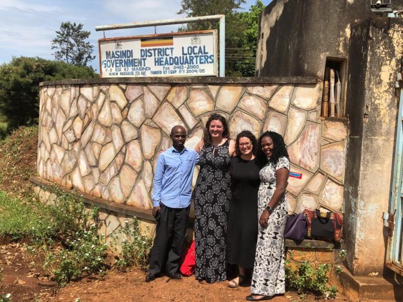 A man in a long sleeve button down light blue shirt standing with three women in long dresses posing outside in front of a stone wall with a rusted rectangular sign on it stating Masindi District Local Government Headquarters some smaller print under with a flag painted at the top of the sign and