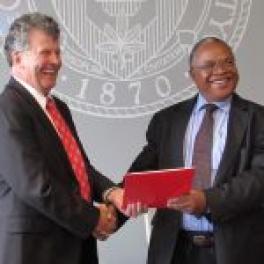 Smiling, two older men, one white, the other black, shake hands and hold a red folder between them