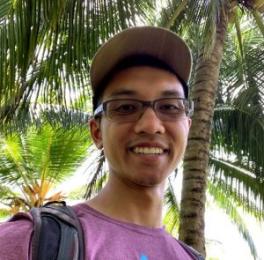 Daniel Ma, a dark-skinned Asian man wearing glasses, a baseball cap, and a backpack. He is standing in a grove of palm trees