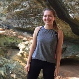 Lillian Ostrander, a white woman with a blonde ponytail wearing athletic clothes and standing in a mossy cave