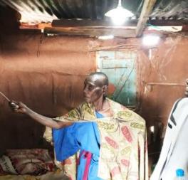 A black person with a shaved head wearing a patterned cape and standing in a tin roofed building