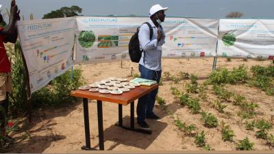 A man with a white hat and long sleeve button down light blue shirt and jeans and a backpack stands in front of a number of banners surrounding him next to a table with plates of some type of bread set out individually outside on Dry land with light soil and small crops with trees in a distance under blue skies