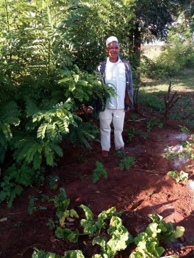 A Black man wearing a white shirt and pants, standing amindst tall plants and new growth