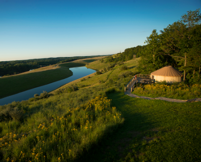 A grass covered hill with a blue river running across it. The sky is clear. On the tall side of the hill is a hut with a fence and a forest behind it