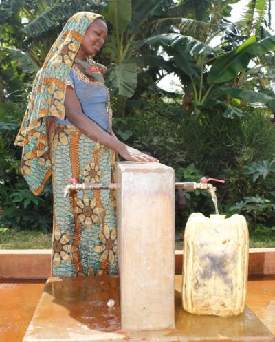 Woman standing next to an outdoor water faucet filling her large yellow canister. 
