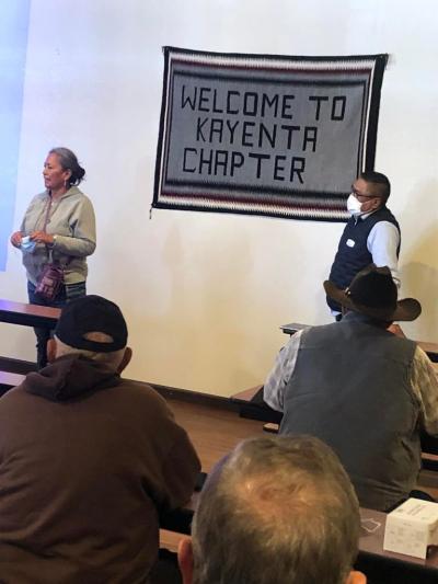 A lady in tan long sleeve and jeans is standing in front of a seated group in a room with a fabric material flag on the wall saying Welcome to Kayneta Chapter. a man with a mask and glasses and a dark vest is standing to the left of the flag and the woman is to the right.