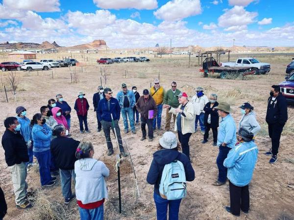 A crowd gathered around a tree planting dig site in a circle as one person is talking to everyone with planted trees sprinkled around and truck and cars parked close by and the mountains at a distance and blue sky with white puff clouds. 