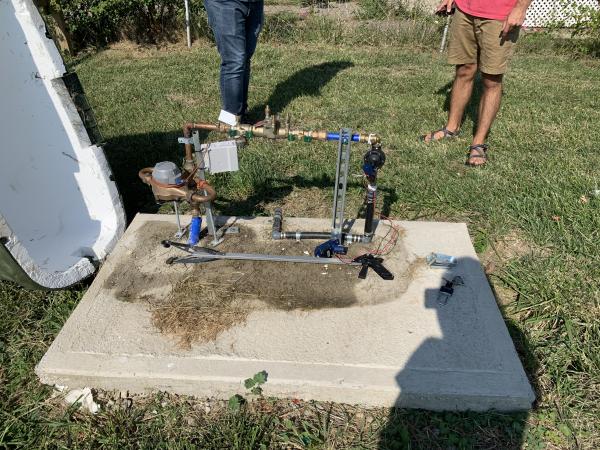 some plumbing contraption on a cement pad in the lawn with the legs of 2 individuals visible, one with jeans and one with tan shorts and a red shirt