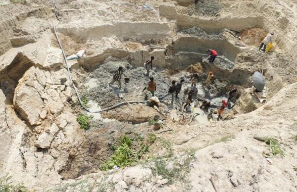 Miners work deep in a pit in Africa. The soil is light colored and there are a few green plants 