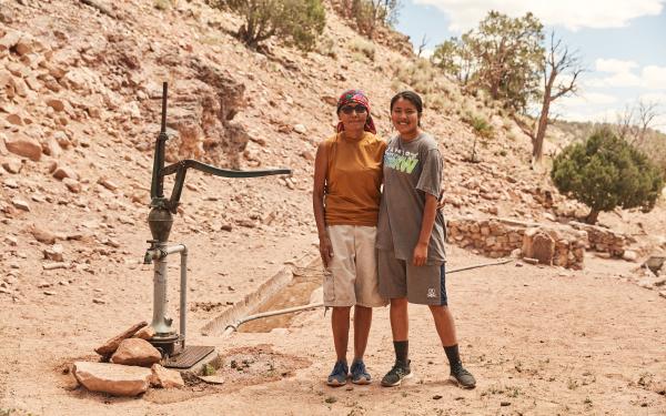 Two people standing next to a water pump posing next to a dirt rocky hill in a desert like environments 