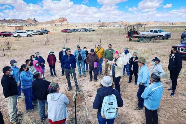A crowd gathered around a tree planting dig site in a circle as one person is talking to everyone with planted trees sprinkled around and truck and cars parked close by and the mountains at a distance and blue sky with white puff clouds. 