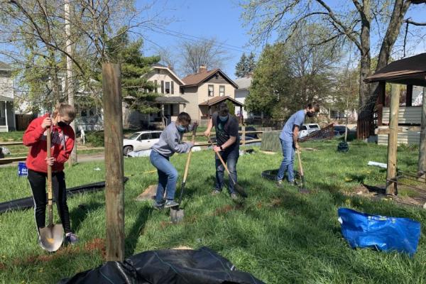 a group of 4 people with shovels digging in the lawn in a neighborhood with houses and trees around and cars in the street behind them under a blue sky.