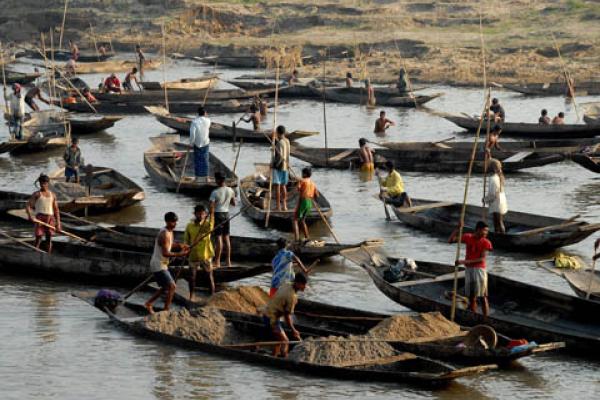 Dozens of shallow boats floating in a river. They are steered using poles. Inside many of the boats are piles of dirt