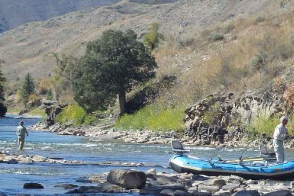 Two people on a river. One is standind in the water, the other is on a rubber, inflatable raft. Both are fishing