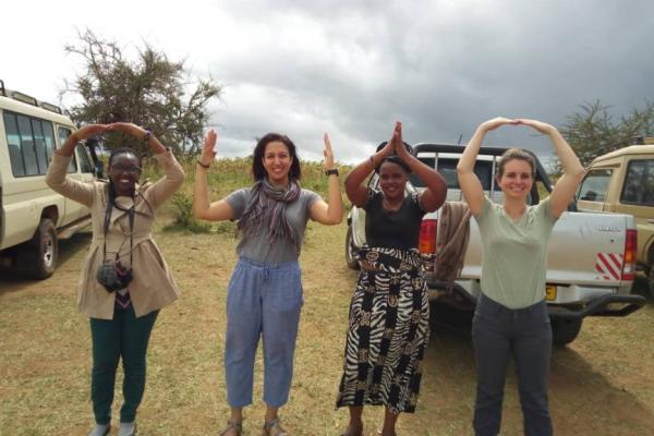 A group of four women making the O-H-I-O shape using their arms. They are standing in an unpaved parking lot with cars behind them