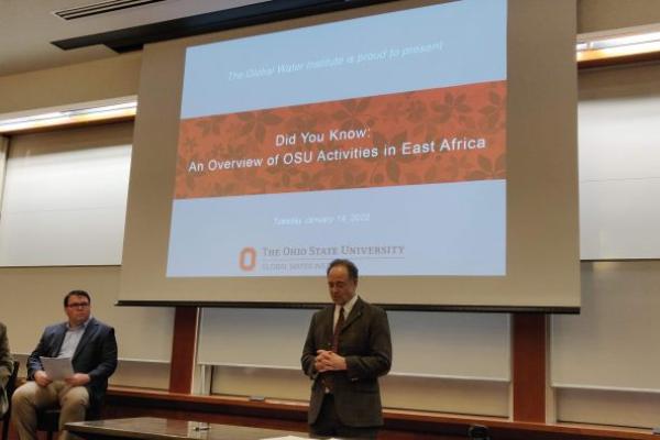 One man standing in front of a projection screen with two men sitting off to the side. Projected onto the screen is a slide reading "Did You Know: an Overview of OSU Activities in East Africa"