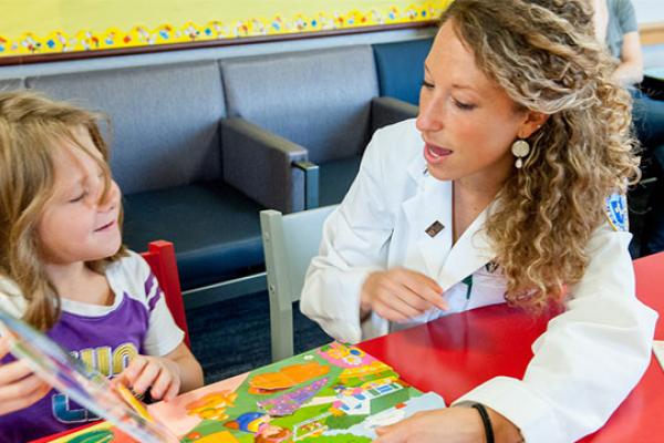 A blonde white woman wearing a lab coat and a blonde white child reading a book together
