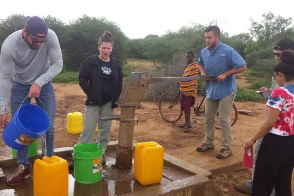 A group pf people using a metal hand pump to gather water into plastic containers