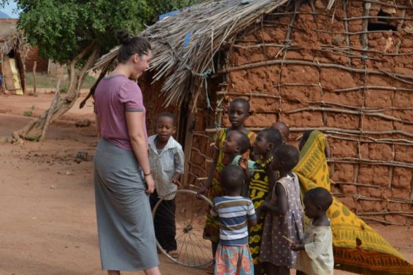 A white woman and a bunch of black kinds smiling and engaged with each other. There is a clay and straw house behind them