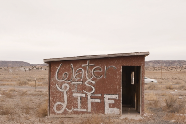 A small shed without a door standing in an empty, dry desert. It is surrounded by bits of yellow brush with low hills and dark shrubs dotting the background. Spray painted onto the shed in curly letters is the phrase: water is life