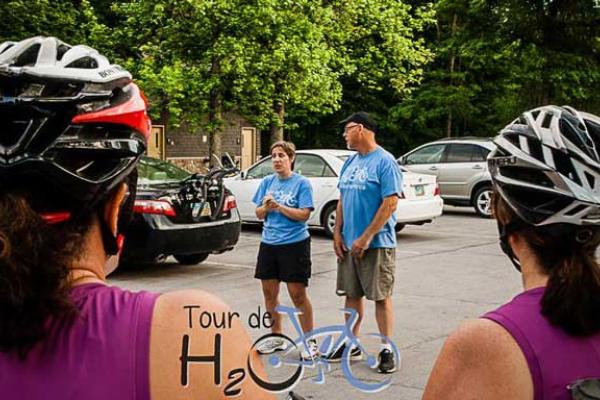 Two white people, a man and a woman, facing the camera and wearing blue "tour de H2O" tee shirts. Facing them are two women in purple tank tops and biking helmets with their backs to the camera.