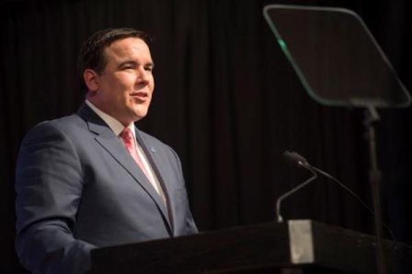 A white man with short brown hair wearing a business suit as he speaks at a podium