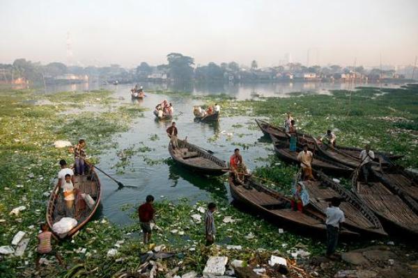 A river full of trash, plants, and boats. There are people in the boats and wading in the water. On the far bank are buildings and trees