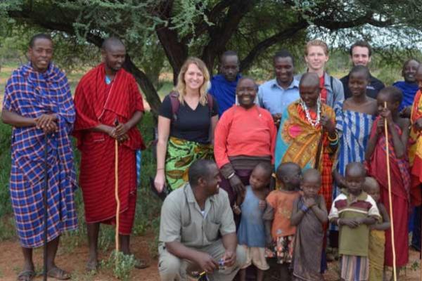 A mixed race, mixed gender group of people standing together beneath a tree and smiling. Many are wearing colorful wraps and patterns.