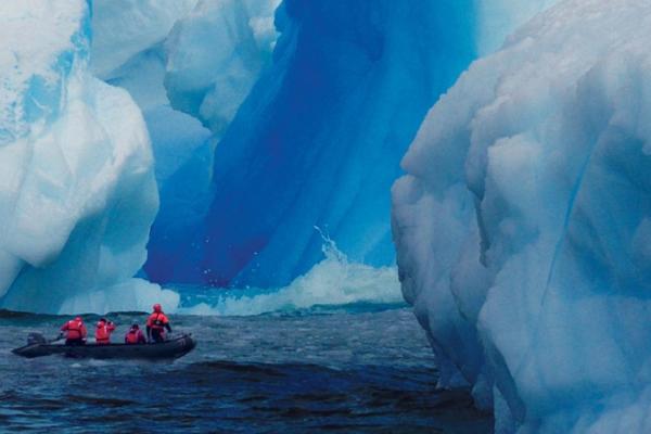 A massive iceberg and a boat with four people in red coats riding across the water towards a break in the ice