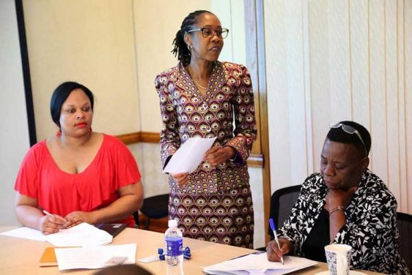 Three Black women in business atire at a meeting table. Two are sitting, the third is standing