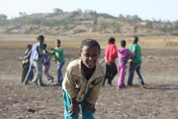 A black boy bent at the waist, hands on his knees, and smiling towards the camera. Behind him, a group of kids are playing