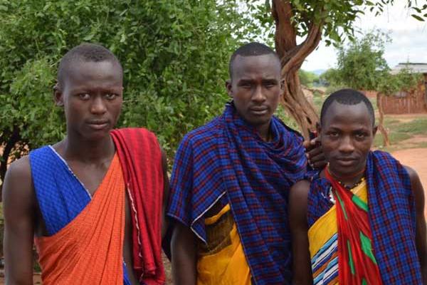 Three black men in brightly colored wraps standing in front of some trees on a red dirt road