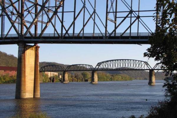 Two bridges crossing over a wide, blue river. The land is lush, green, and hilly. There are nearby buildings on the far side of the river.