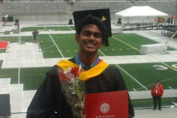 An Indian man with dark skin and black hair smiling at the camera. He is wearing a graduation cap and gown, and holding a bouquet of roses and a diploma