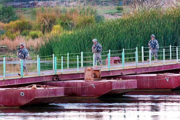 Three armed soldiers in combat gear walking across a bridge over a river