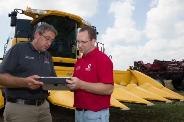 Two white men wearing Ohio State shirts. They are holding an iPad between them and standing in from of a large, yellow machine