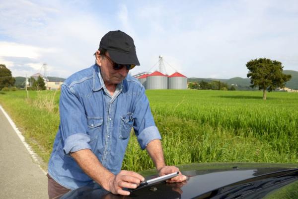 An older white man in a black cap and jean shirt. He is leaning over the hood of a car and looking at a tablet
