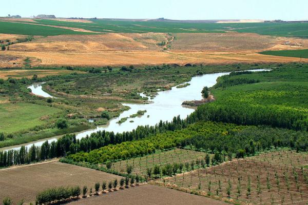 A river flowing between fields and trees and a broad countryside with a few buildings at the peak of the hills