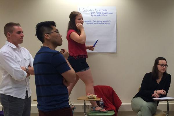 Four students, two men and two women, looking at something off camera. The men are standing with their arms crossed. One of the women is seated. The other is standing between two chairs with a marker in hand and a large sheet of paper on the wall behind her, ready to write more.