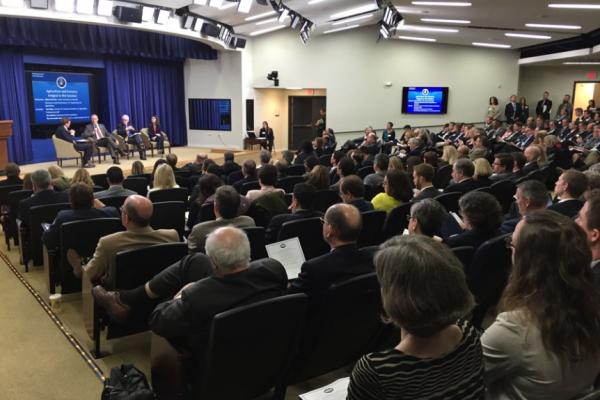 An full auditorum watching four people in chairs on stage. Behind the people on stage is a presentation slide.