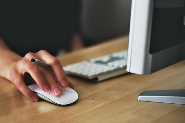 person's hand holding a pen and using mouse on the desk with a keyboard and monitor,