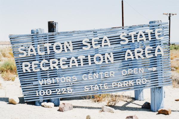 A Blue wooden sign with white letters outdoors says Salton Sea State Recreation Area Visitors Center Open 100-225 State Park Rd.