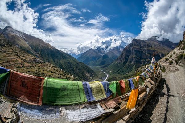 Snow capped mountains in a distance with green mountains int he foreground with a valley below and a river running through it. There are prayer flags hanging in the foreground along a dirt road.