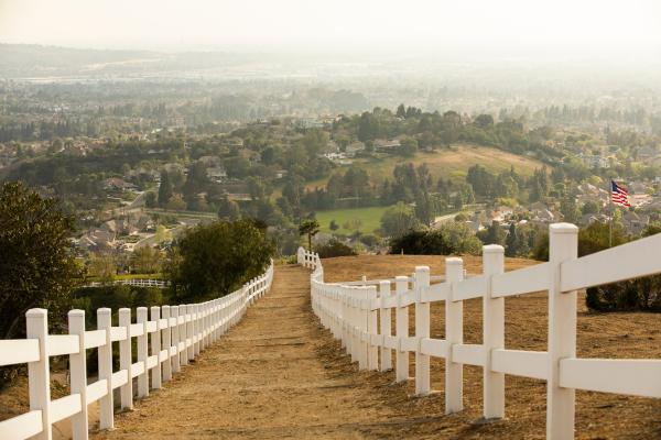 A view of the hazy city from a hill top from a dirt trail with a white picket fence on either side.