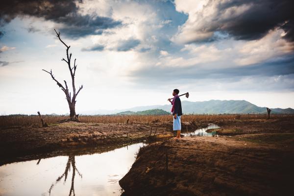 Farmer standing with a tool in arid land next to a water stream with dry vegetation around and green mountains in a distance under dark cloudy skies