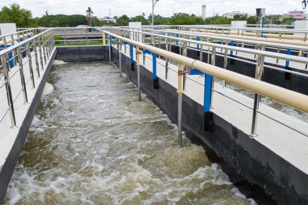 Water flows through a large outdoor water treatment facility with numerous metal guardrails and concrete structures, under a partly cloudy sky.